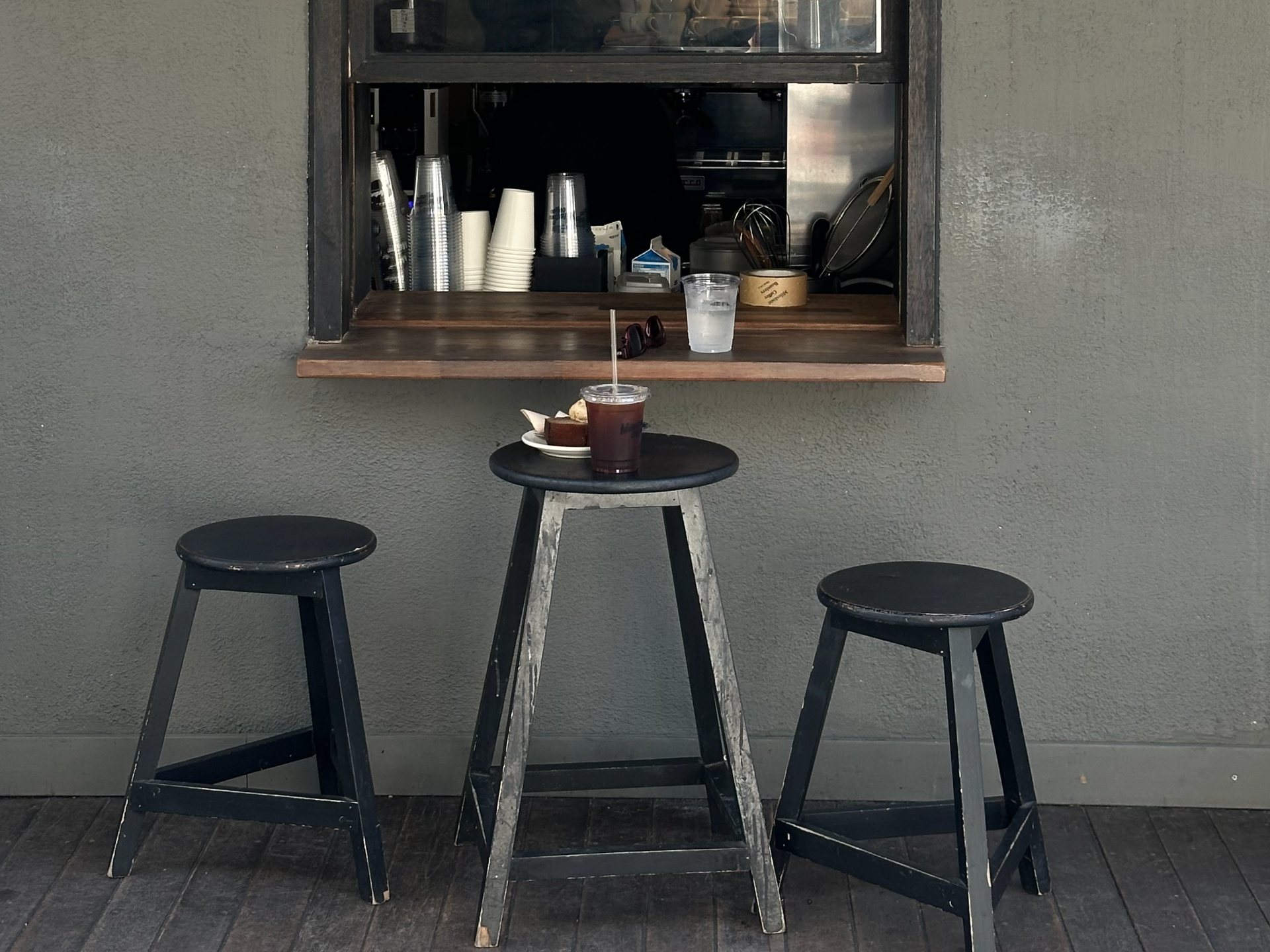 Coffee-shop service window with a wooden ledge, three black stools, and a stone-grey exterior wall