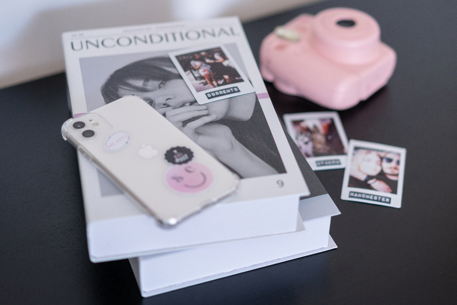 Flatlay of a black-and-white magazine, a clear-cased smartphone, a pink instant camera, and polaroid prints on a dark table