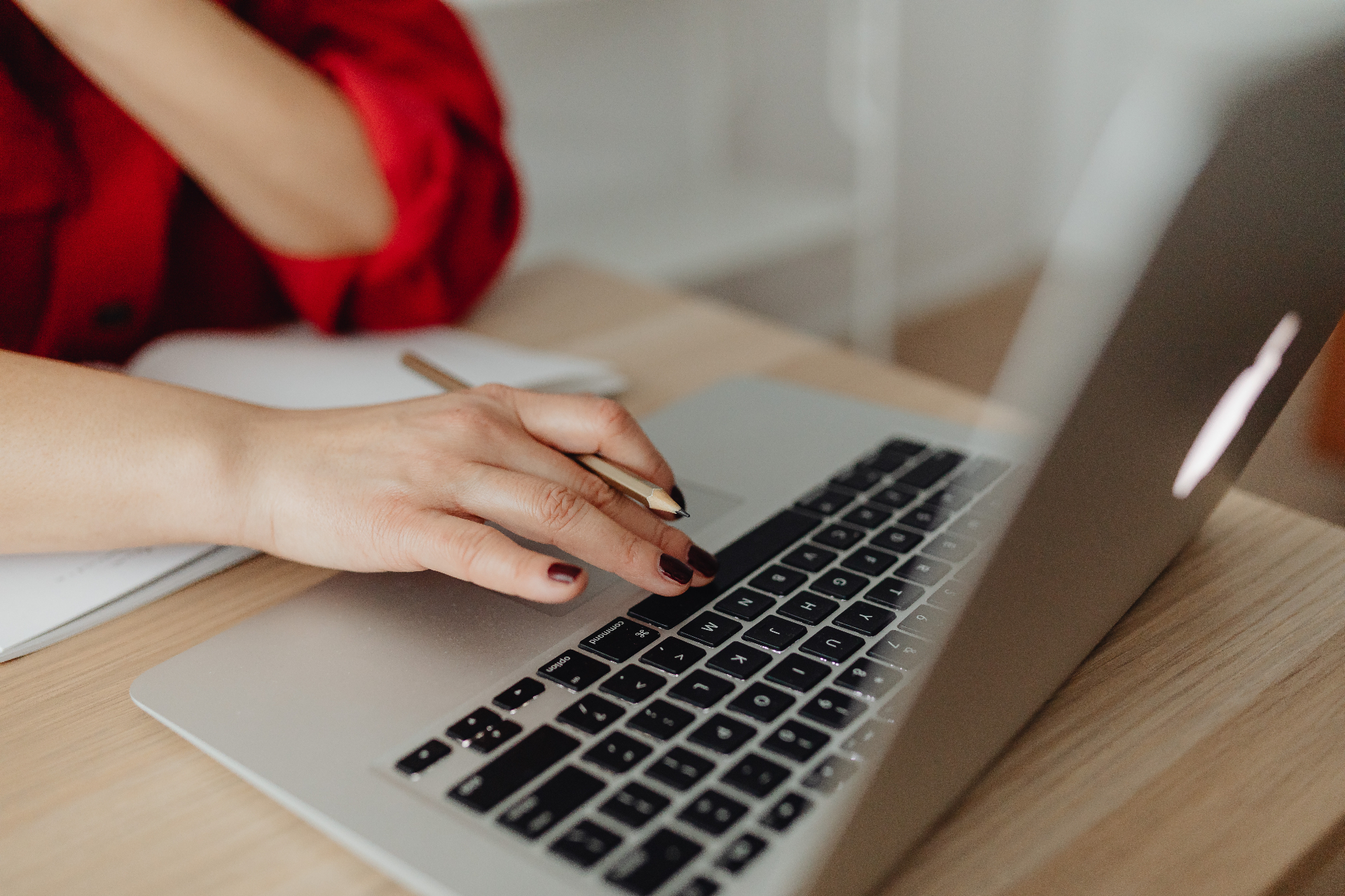 Woman in a red top typing on a silver laptop at a light wooden desk, holding a pencil