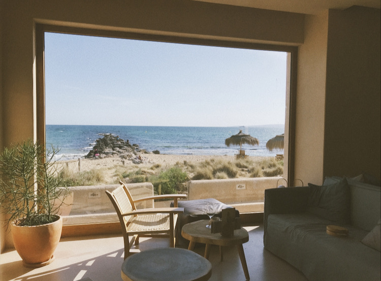 Sunlit coastal living room with a rattan chair and large picture window overlooking a sandy beach and calm ocean